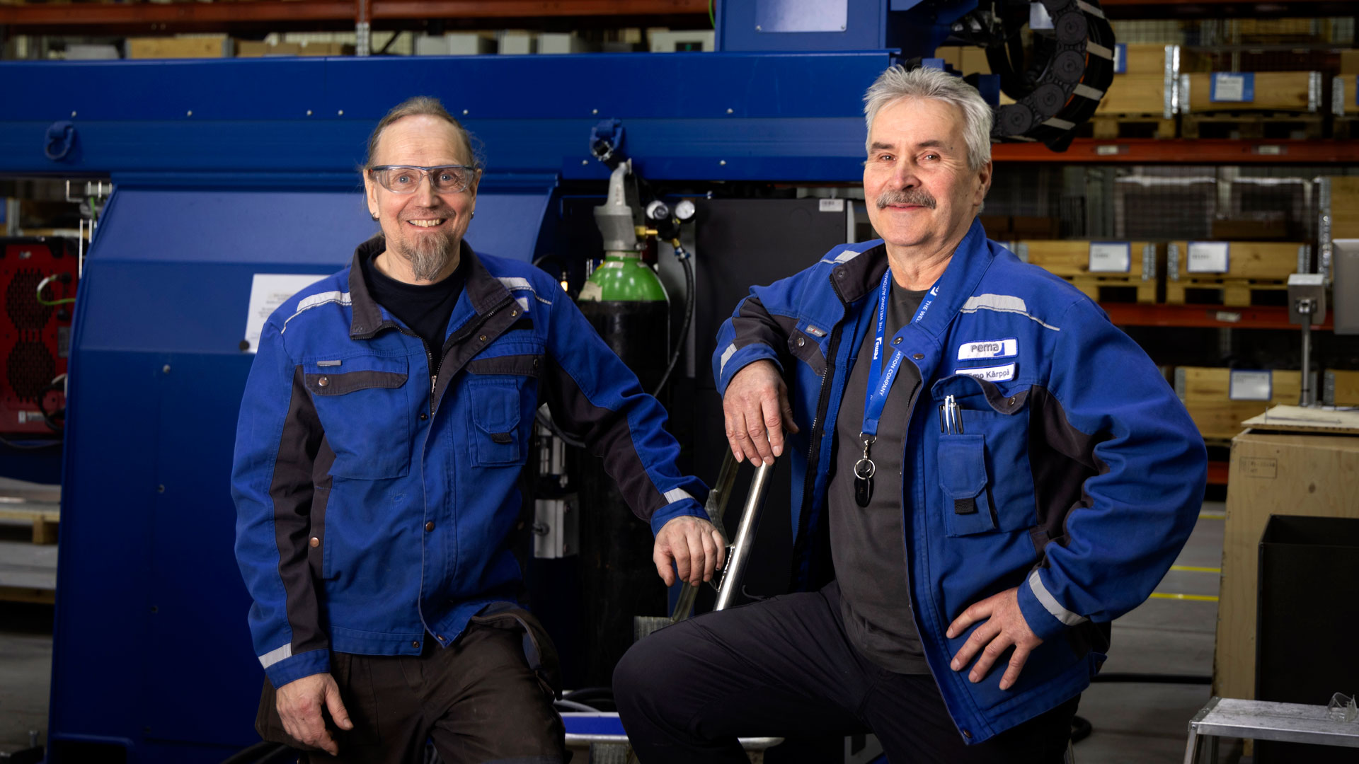Kaksi miestä sinisessä työtakissa hymyilee työkoneen edessä.