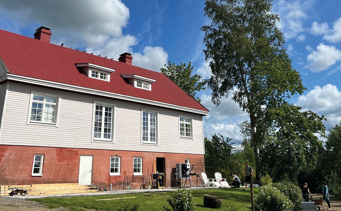 Kuvassa vaalea puutalo, terrakotan värinen kivijalka ja tummanoranssi katto. Pihalla muutama ihminen, puita ja nurmikkoa.