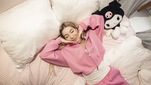 Nelli Orell makaa sängyllä hymyillen ja pitelee kämmenselkiään leukaansa vasten silmät kiinni, vieressä on valkoinen nalle mustassa lakissa.