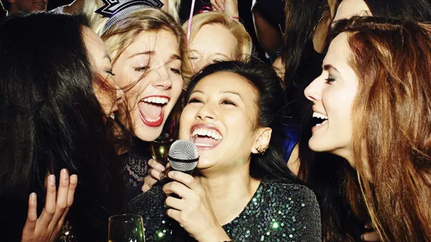 Viisi naista laulaa samaan mikrofoniin, mikrofonia pitelevä nainen hymyilee.