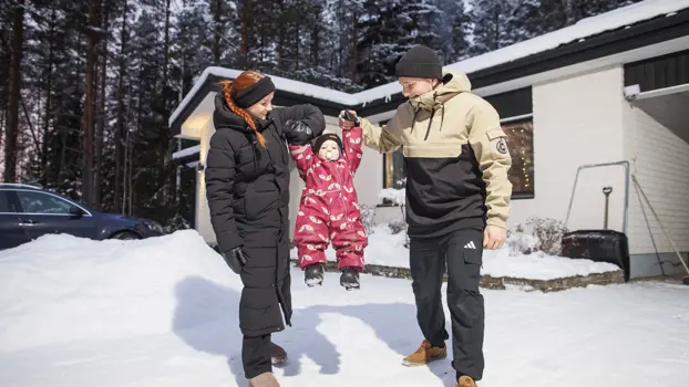 Inka Keteli ja Otto Ikonen roikuttavat hymyillen ilmassa lastaan Eleaa omakotitalonsa edessä talvivaatteissa. Maassa on lunta.