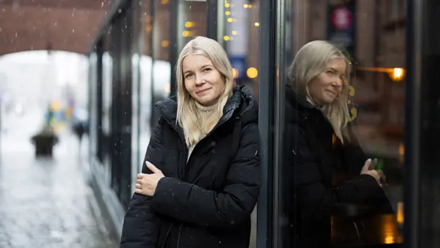 Tinja Tähtinen katsoo kameraan ja nojaa lasiseinään yllään musta toppatakki, taivaalta sataa lunta.