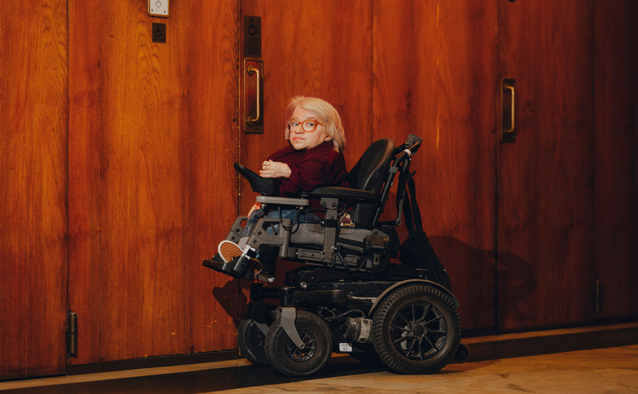 Anni Kyröläinen istuu pyörätuolissa hissien edessä ja katsoo kameraan.