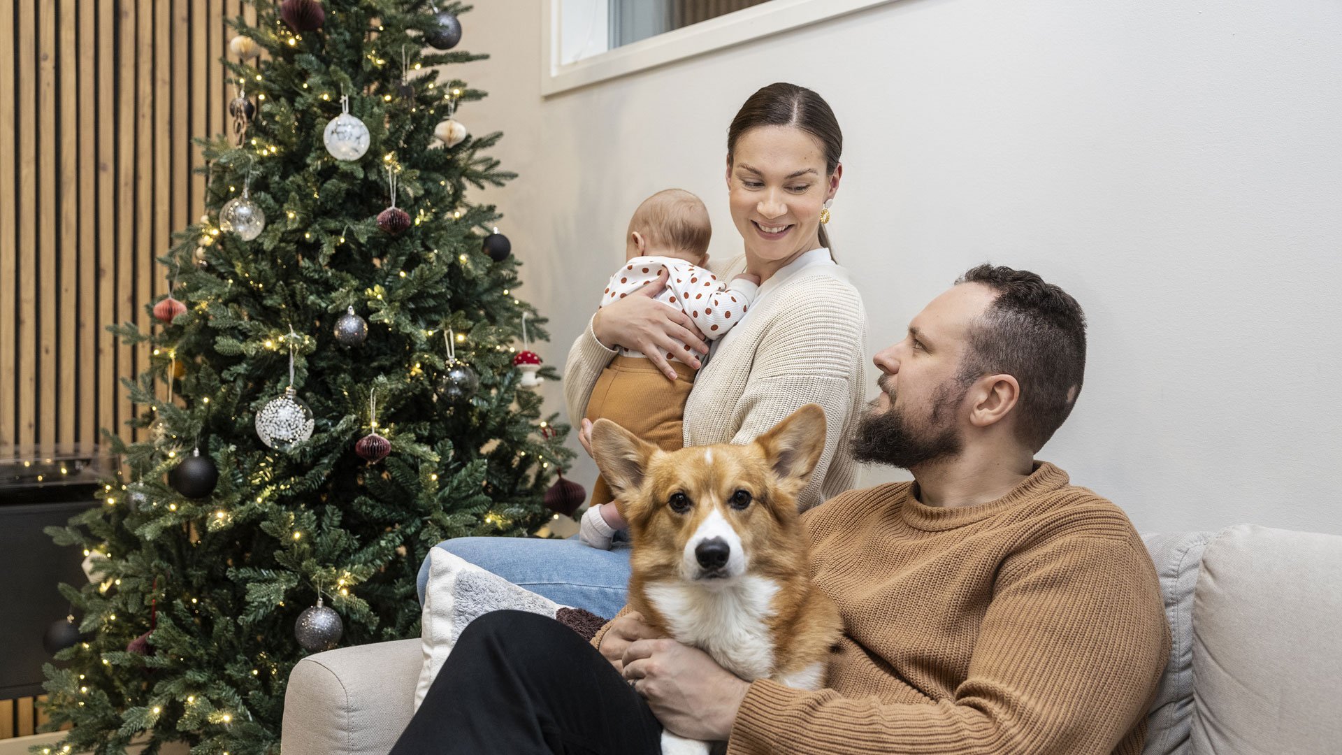 Riina Periviita pitelee lasta sylissään, vieressä Antti Periviita sylissään Manu-corgi. Taustalla näkyy joulukuusi.