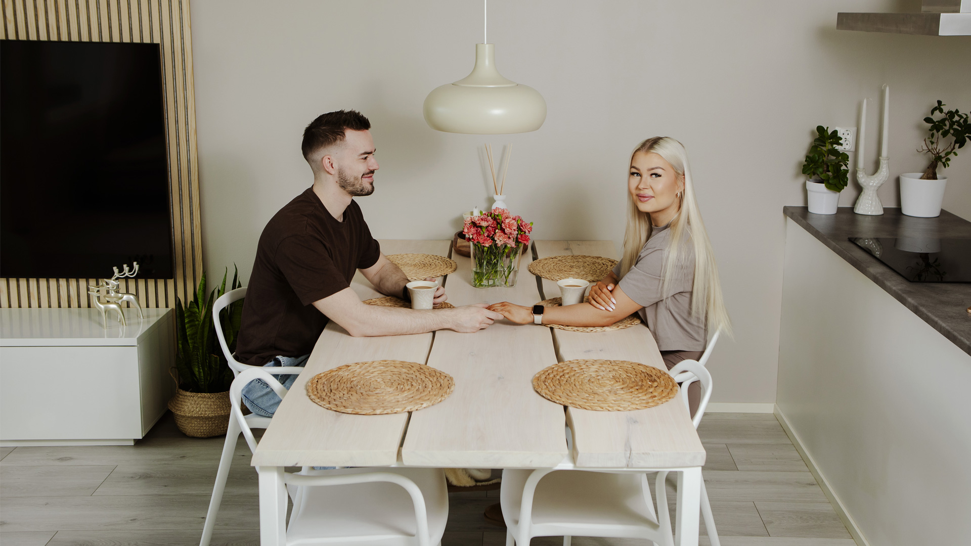 Nuori valkoinen mies ja nainen pitävät toisiaan kädestä pöydän päällä hymyillen nainen katsoo kameraan.