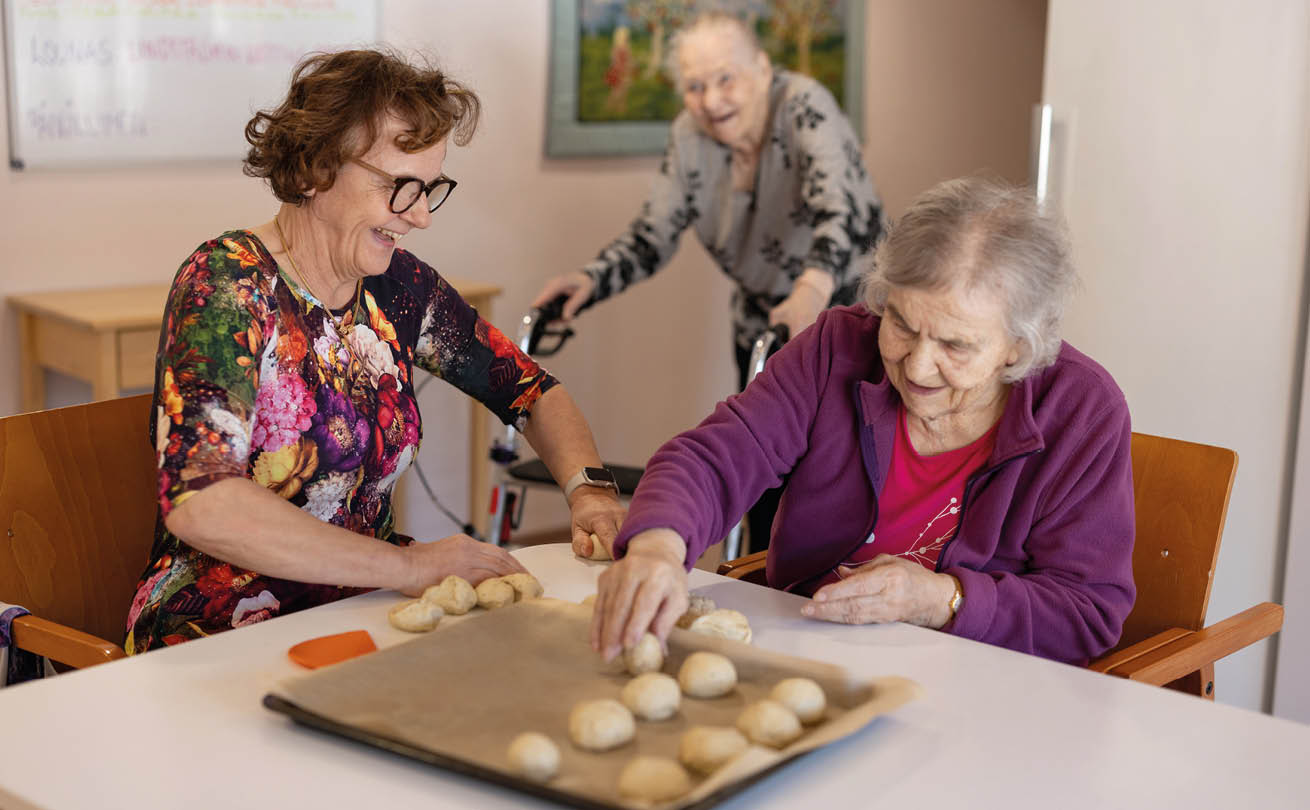 Auli Härkälä leipomassa pullaa Meeri Mahlamäen kanssa. Taustalla Helmi Viljanen
