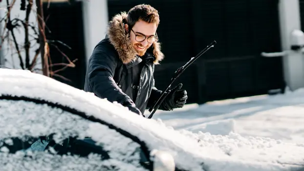 Mies puhdistaa lumista autoa. Auton kylmäkäynnistys, auton käynnistys pakkasella, kuvituskuva