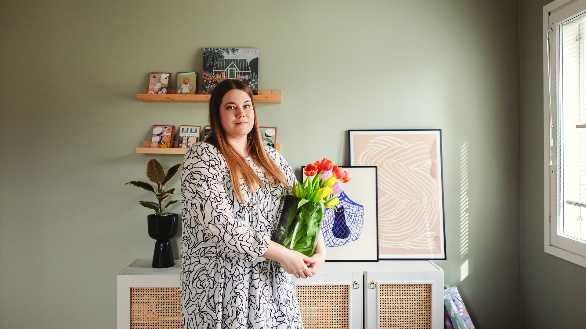 Kuvittaja Kaisu Sandberg seisoo tulppaaniruukku kädessä huoneessa.