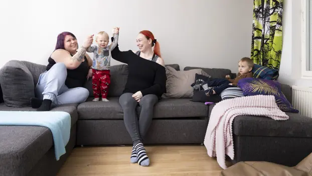 Jenni Tamminen nostaa sohvalla lapsensa Eemilin käsiä yhdessä lasten kummitädin Jennan kanssa. Sohvannurkassa istuu vanhempi lapsi Oliver.