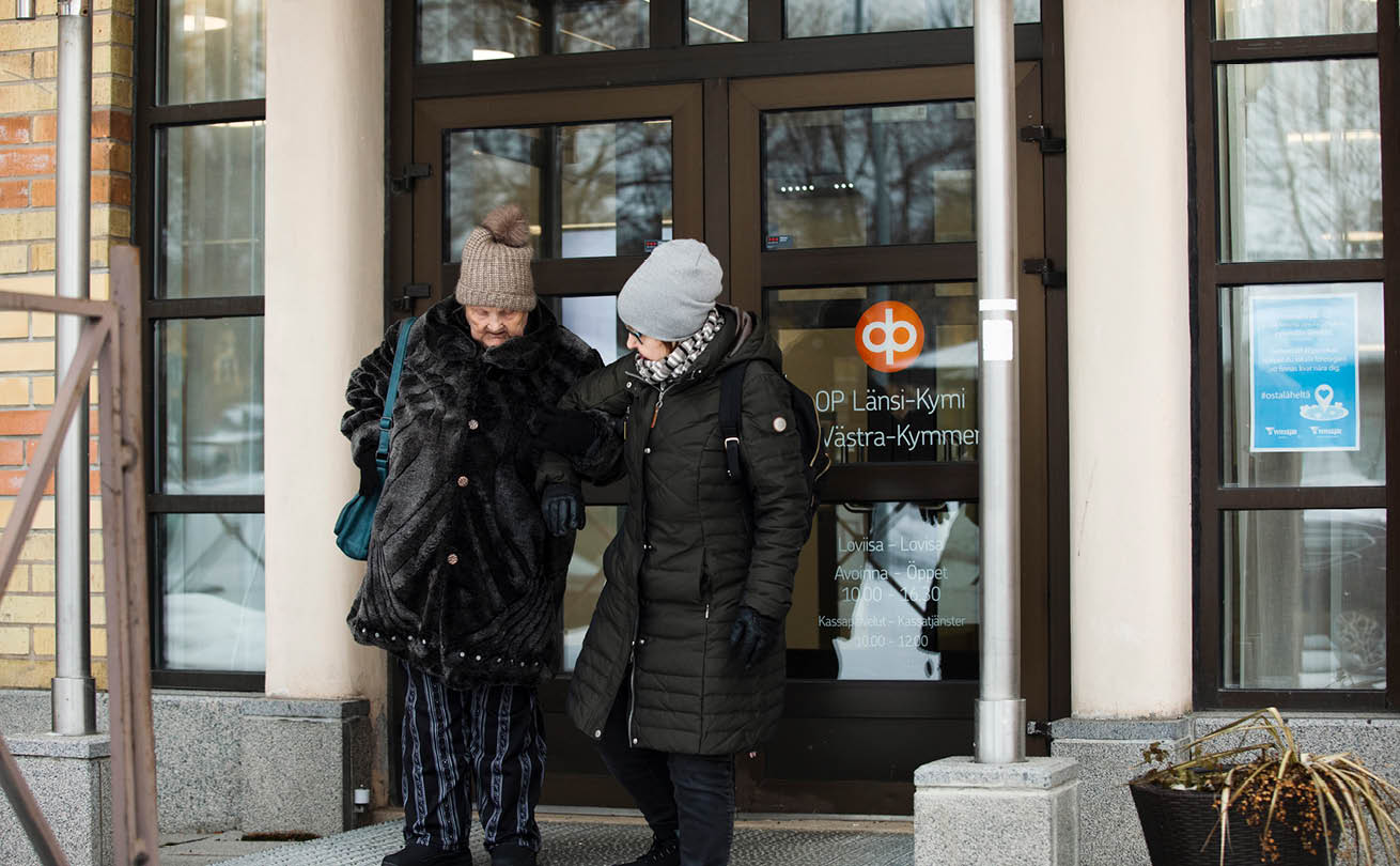 Kaksi naista pankin portailla talvella pipot päässä. Nuorempi auttaa vanhempaa.