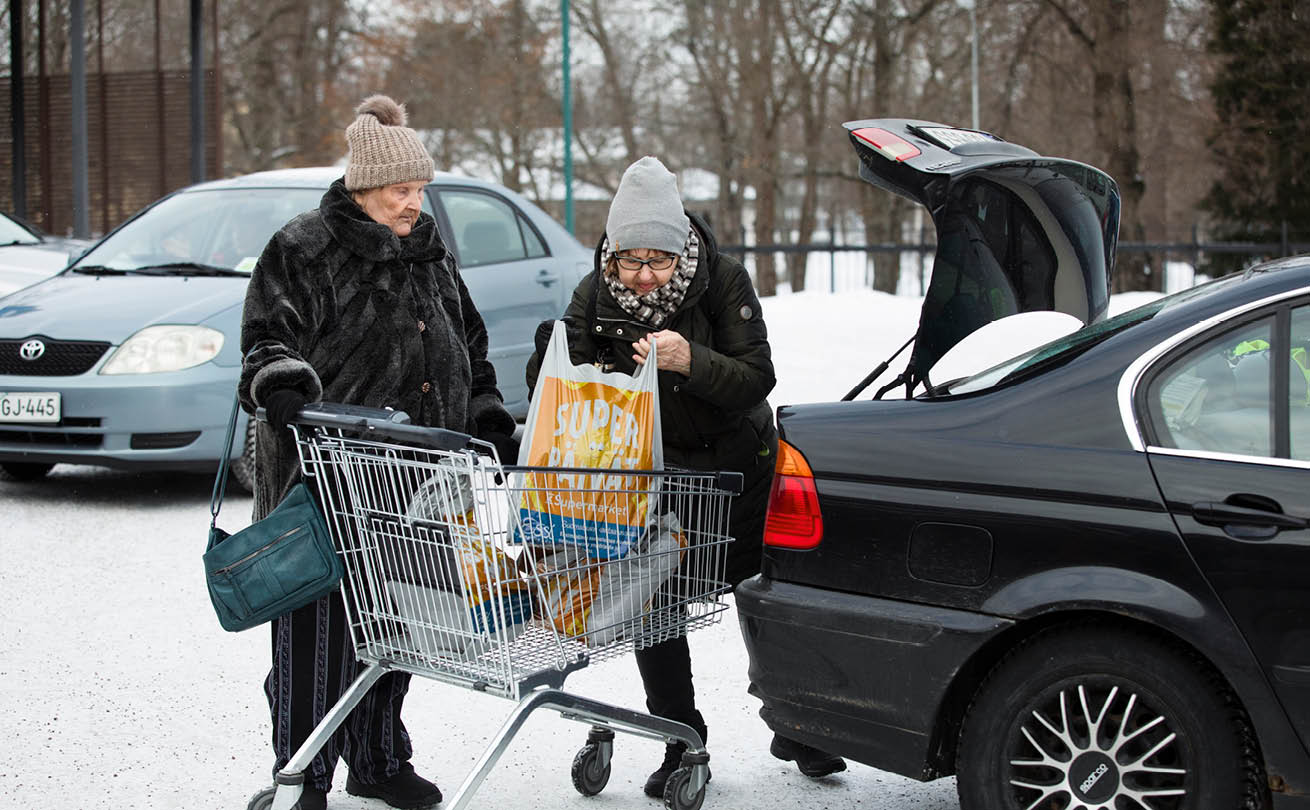 Vanhempi nainen pitelee ostoskärryä, kun nuorempi pakkaa ruokaostoksia auton takakonttiin.
