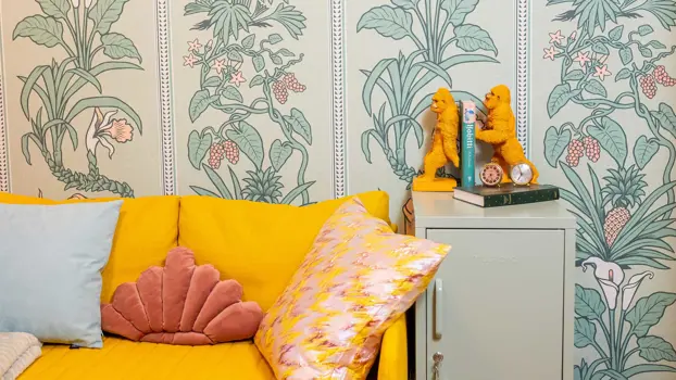 Keltaiset apinafiguurit pitelevät kirjoja pystyssä pienen kaapin päällä, joka seisoo sinapinkeltaisen sohvan vieressä. Vihreänsävyinen tapetti on kukikas.