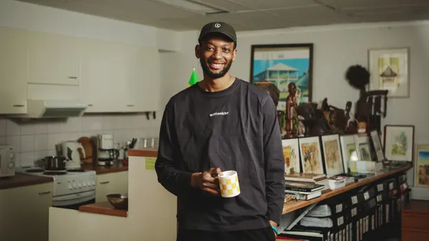 Nosh A Lody seisoo kahvikuppi kädessä ja hymyilee, taustalla näkyy keittiö ja taso, jonka päällä on kirjoja ja taidetta.