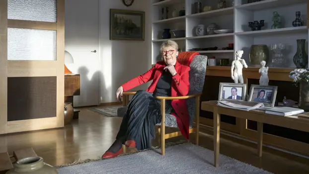 Sirkka-Liisa Kivelä istuu nojatuolissa vieressään pöytä, jolla seisoo kaksi valkoista patsasta. Taustalla näkyy kirjahylly, jonka hyllyillä on astioita.