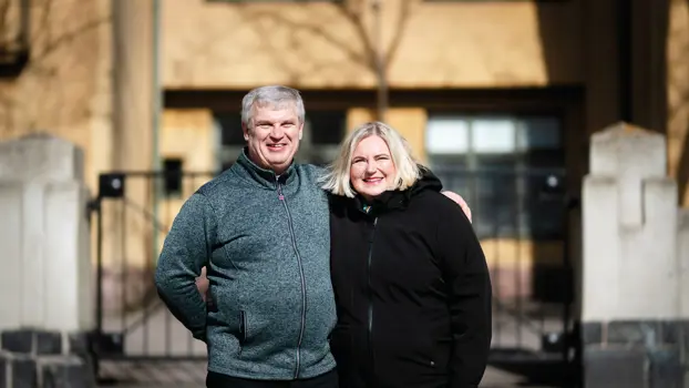 Paula on Ossin kainalossa, ja he hymyilevät, taustalla näkyy keltainen talo.