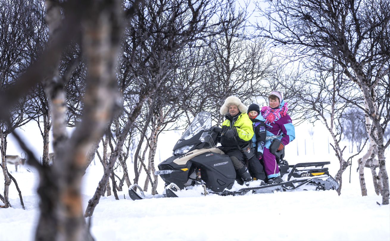 Moottorikelkan päällä kelta-mustassa suojapuvussa mies, takana lapsi ja hänen takanaan nainen turkoosi-magentan värisessä suojapuvussa.