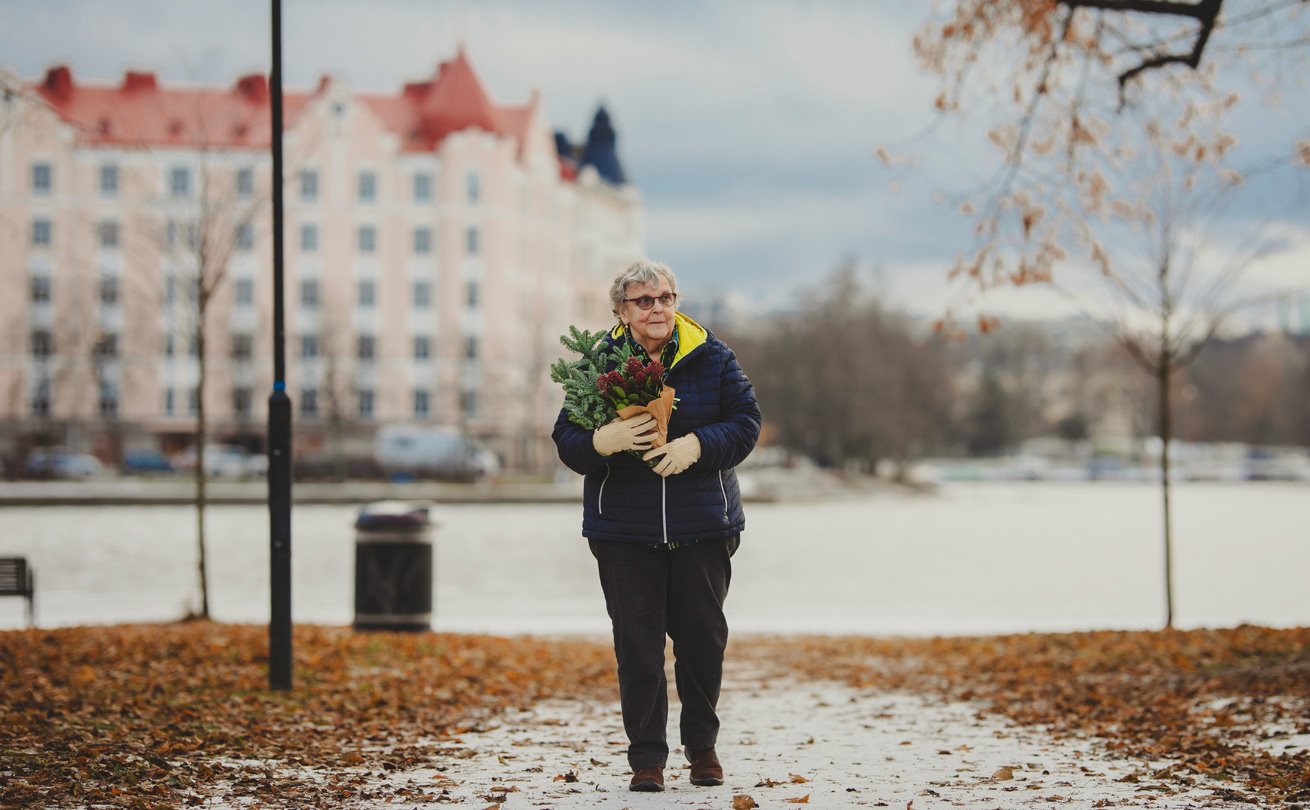 Varttunut nainen kävelee merenrannalla, sylissään havuja ja joulukukka, taustalla vastarannan kerrostalot.