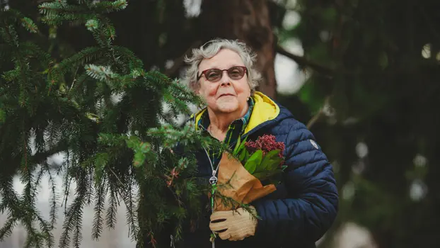 Varttunut nainen pitää sylissään joulukukkaa, toppatakki päällä, taustalla havupuu.