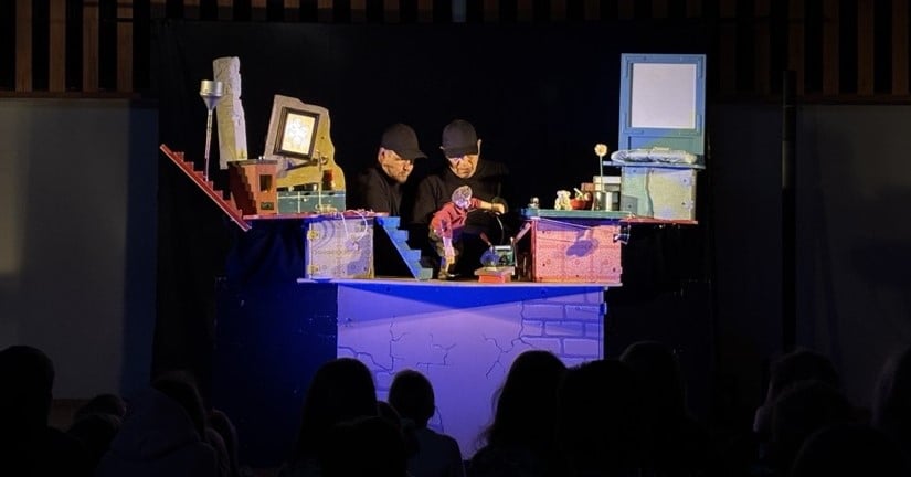 Kuvassa pimeä liikuntasali, jonka seinustalla Poika ja korppi -nukketeatteriesityksen lavasteet. Lavalla esiintymässä Antti Kemppainen ja Hannu Räisä.