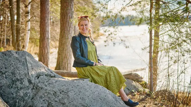 Sisko Viikki-Dufva istuu penkillä vihreässä mekossa puiden keskellä ja hymyilee. Taustalla on järvimaisema.