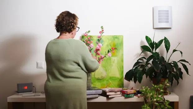 Mirja seisoo selin kameraan ja asettelee vaaleanpunaisia kukkia maljakkoon. Taustalla näkyy tason päällä vihreä taulu ja viherkasveja, kirjoja ja rasioita.