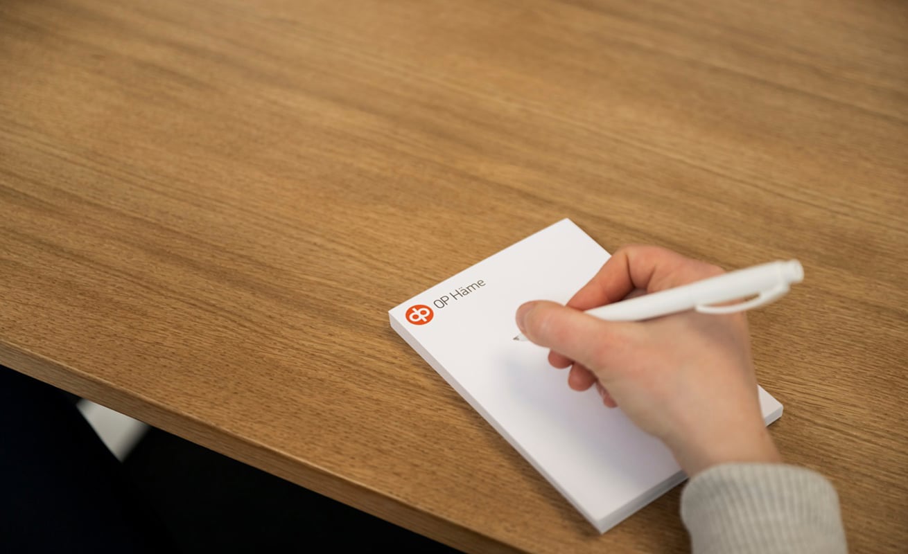 Käsi kirjoittaa muistilehtiöön puupintaisen pöydän ääressä. Muistilehtiössä näkyy OP Hämeen logo, ja kuvassa korostuvat muistiinpanojen tekeminen, suunnittelu ja asiantuntijatyö.