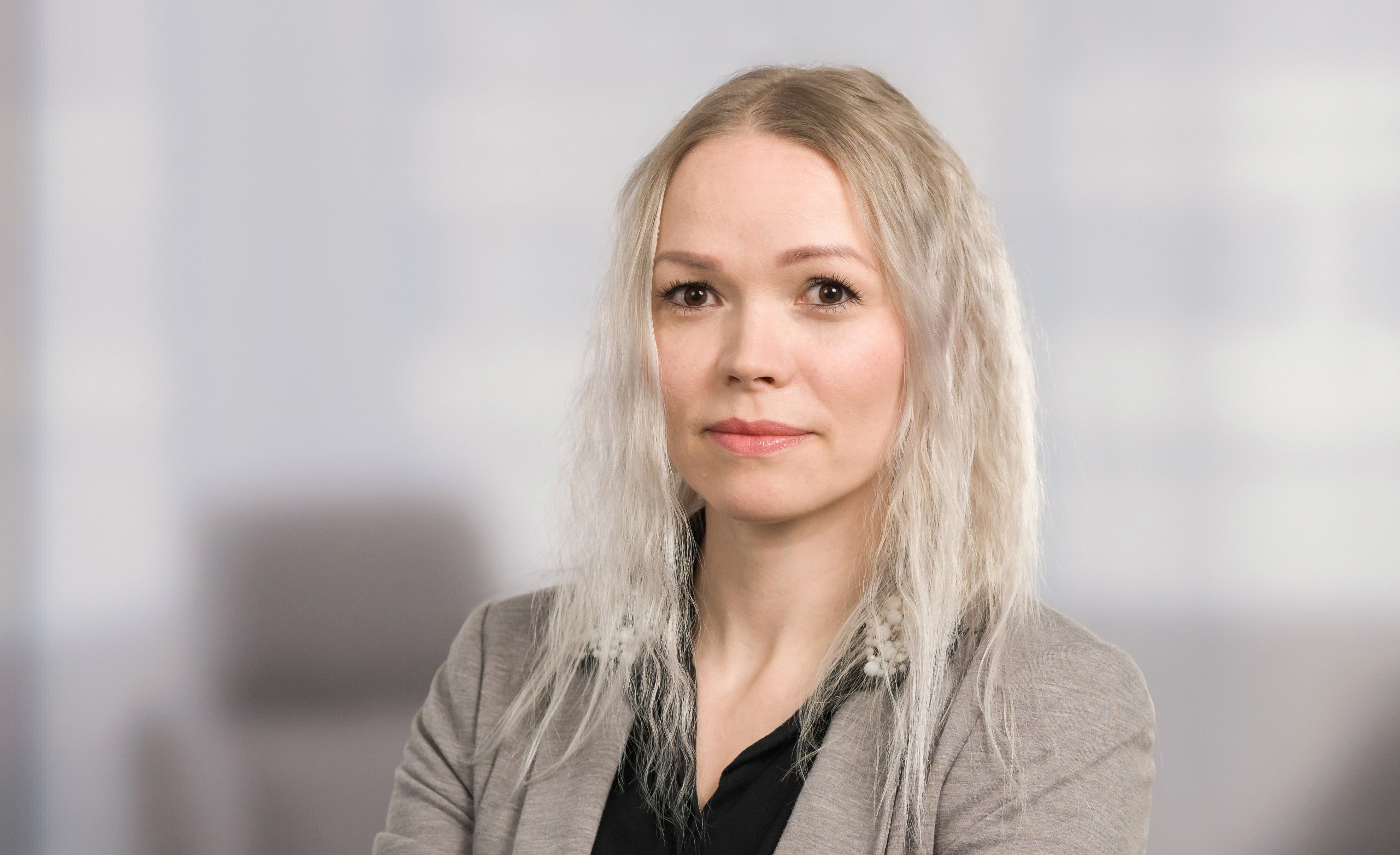 Asiakkuusasiantuntija Stella Hints seisoo toimistoympäristössä kädet ristittyinä. Stella Hintsilla on vaalea bleiseri tumman yläosan päällä, ja taustalla näkyy pehmeästi sumentunut toimistotila. Kuva on ammatillinen henkilökuva, joka korostaa asiantuntijuutta ja lähestyttävyyttä.