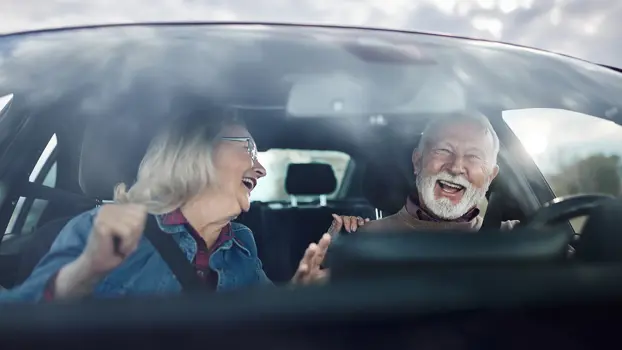 Charmantti vaaleatukkainen mies ajaa autoa. Puoliso hymyilee hänelle matkustajan paikalta.