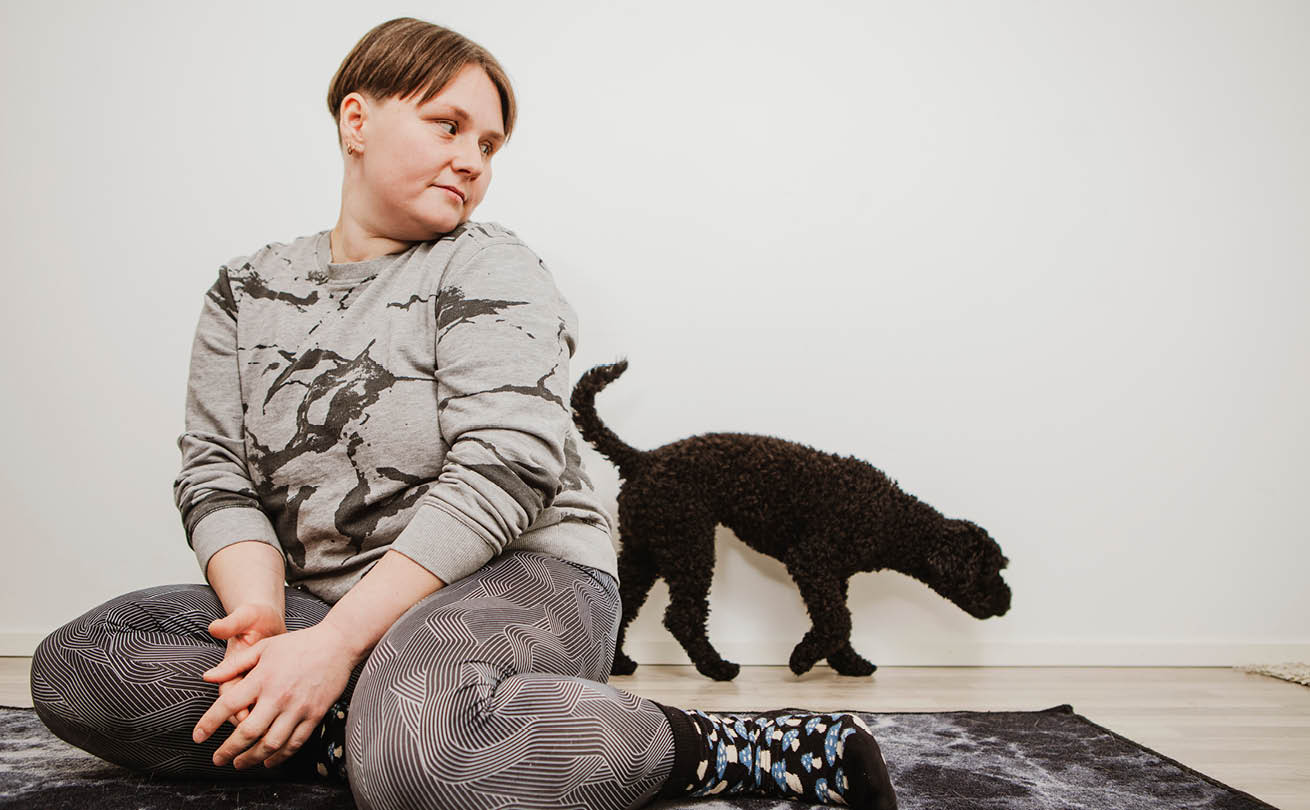 Nainen istuu lattialla ja katsoo mustaa koiraa, joka on hänen takanaan.