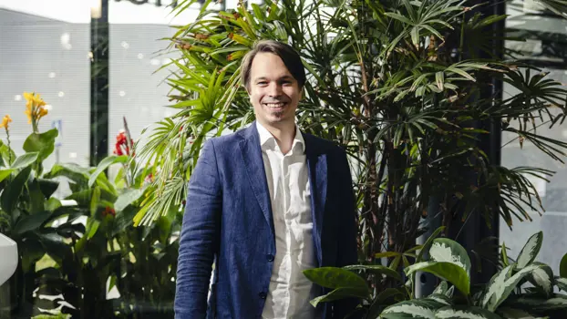 Themo Nordicin Markus Perkkiö hymyilee iloisesti. Päällään hänellä on sininen rento pikkutakki. Taustalla näkyy isoja viherkasveja.